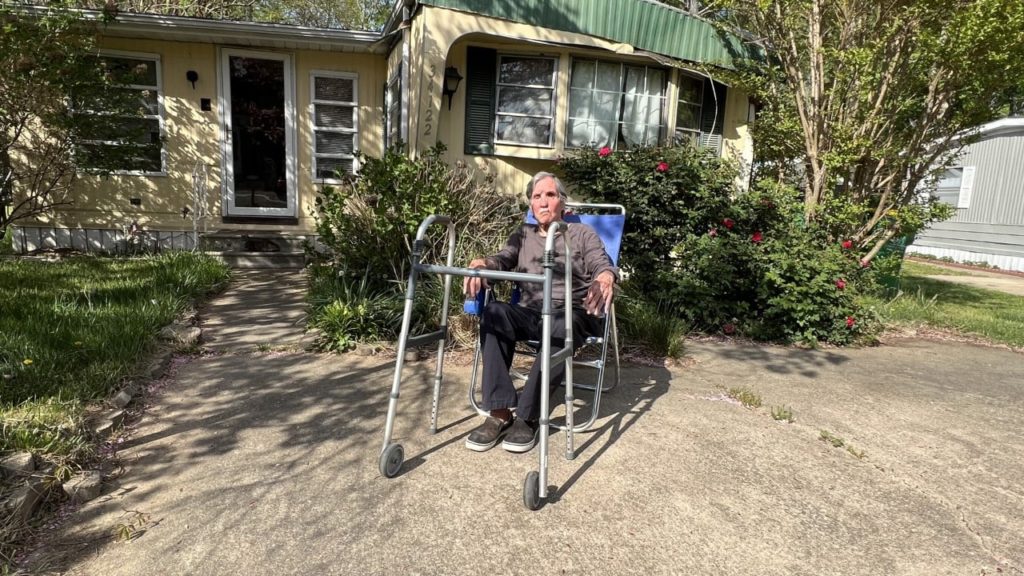 Robert Kravis seated in a lawn chair with a walker in front of him outside.